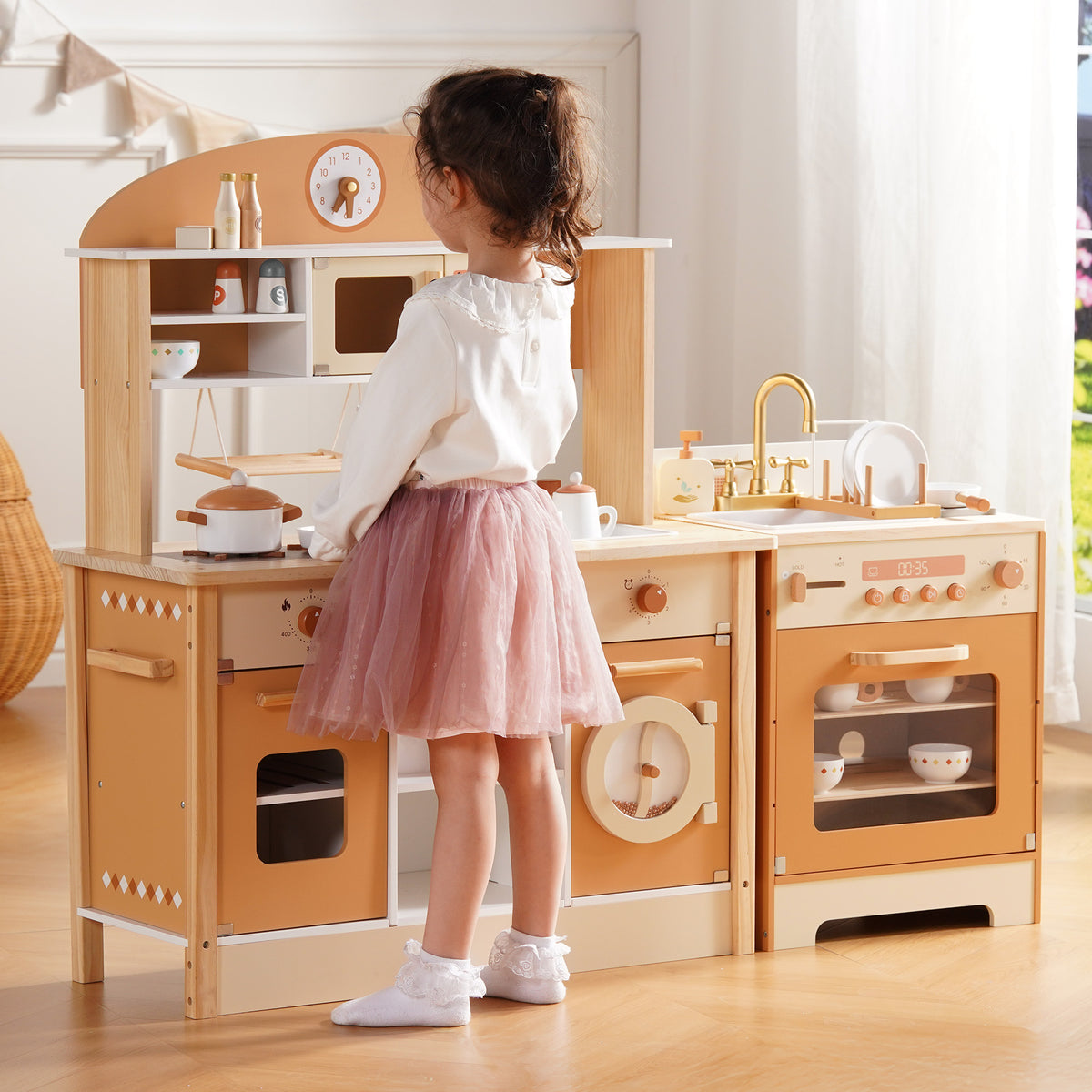 Young girl in pink tutu skirt standing at ROBUD Caramel Wooden Kitchen playing with pots and brass-accented stovetop