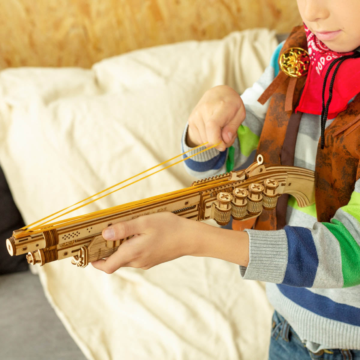 Child in striped shirt aiming a ROKR wooden rifle model sideways in a workshop, wearing brown vest and red bandana