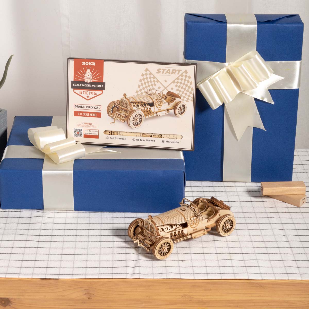 ROKR Grand Prix wooden car model beside retail box and blue gift boxes on white grid tablecloth