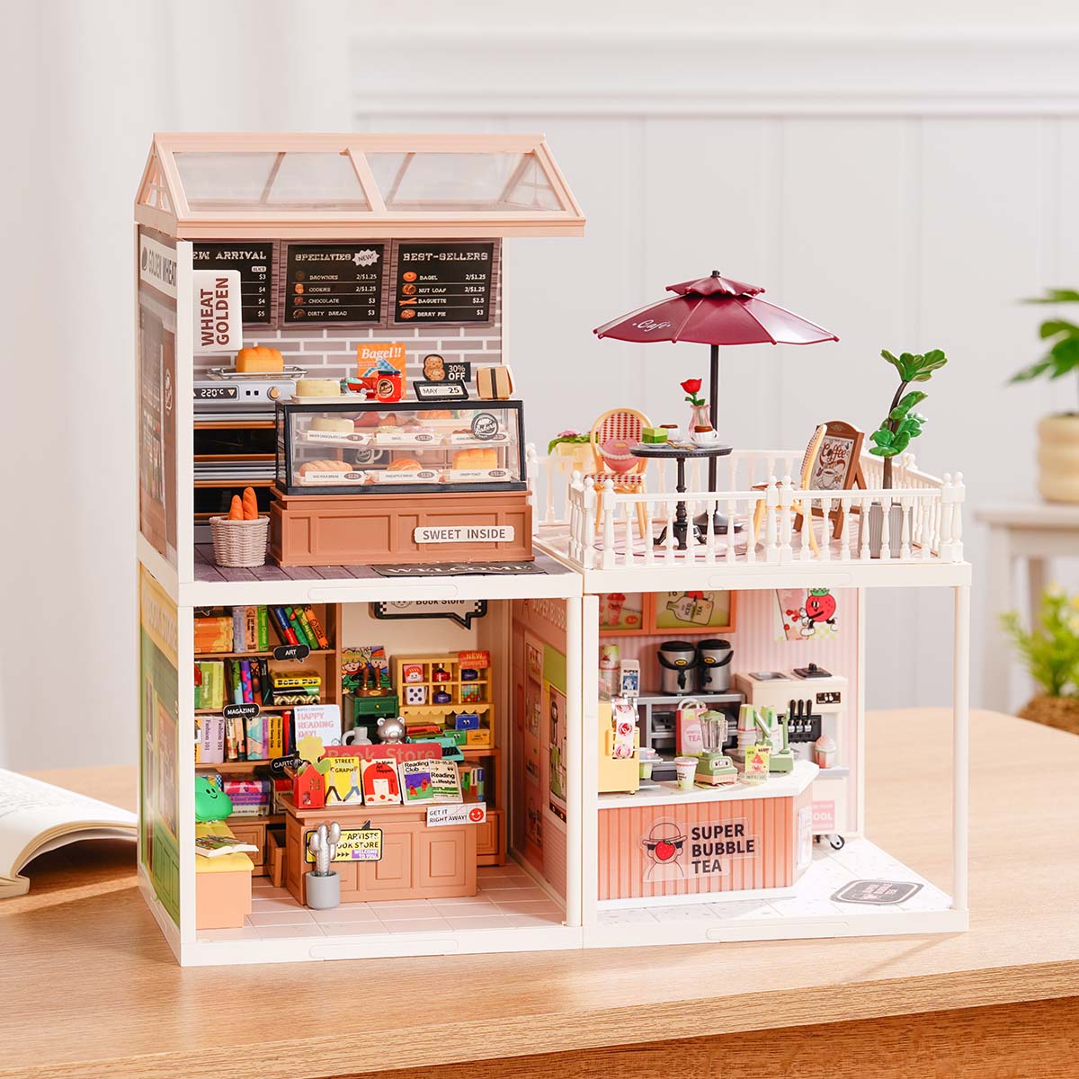 Rolife Super Creator Series 3-in-1 L Shape front view showing a coffee shop and a fruit store in the lower L frame plus a rooftop balcony with sunflowers on a wooden desk beside a plant.