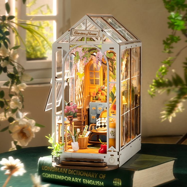 Rolife Garden House book nook with warmly lit miniature interior behind glass walls, resting on stacked hardcover books
