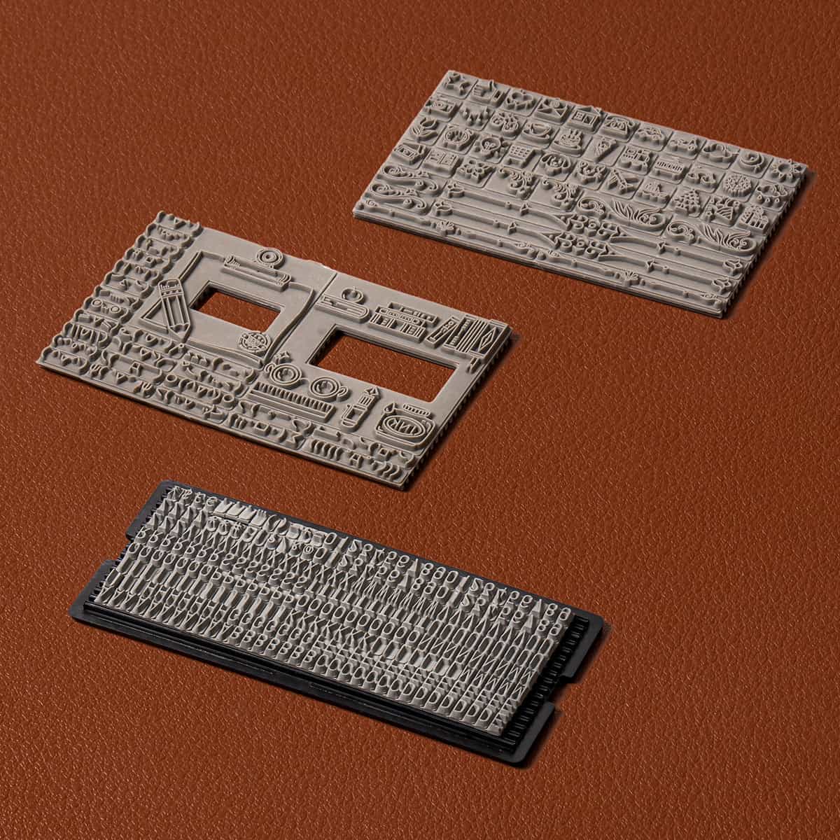 Three ROKR Classic Printing Press moveable type blocks with engraved text characters on a rust brown leather surface
