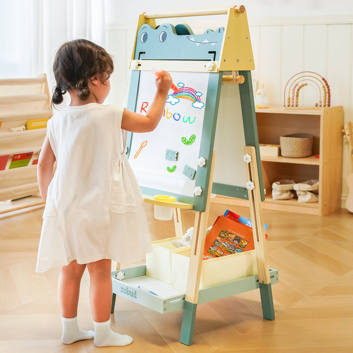 Toddler girl drawing on the ROBUD Wooden Crocodile Easel whiteboard side in a bright playroom