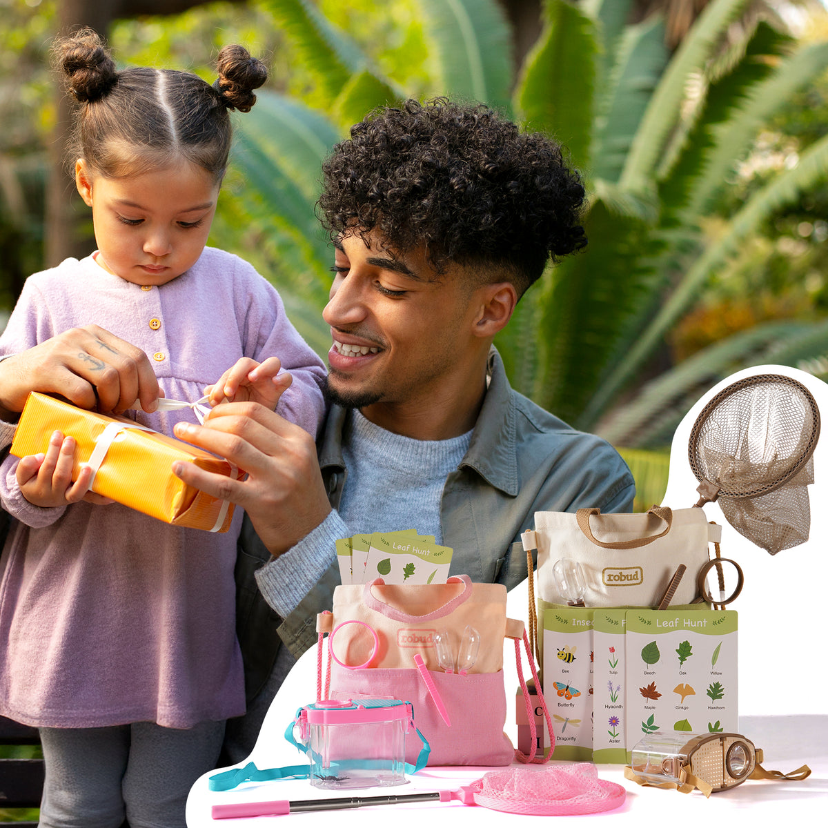 Two girls in meadow sitting on grass playing with ROBUD pink and green outdoor explorer kit accessories in warm golden afternoon light
