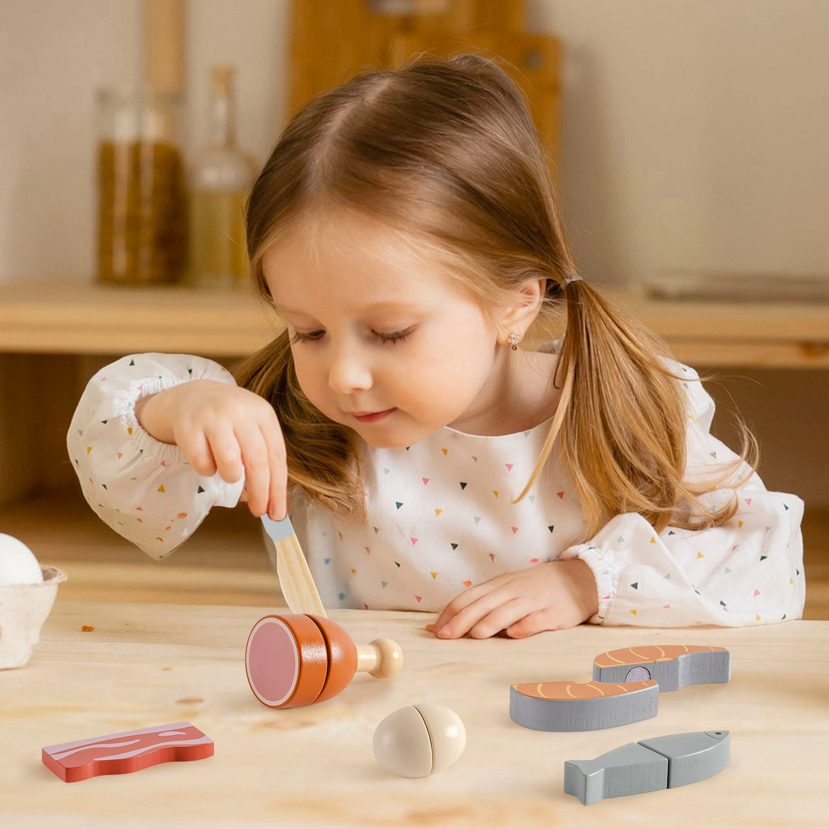 Young girl cutting a wooden chicken leg piece from the ROBUD meat playset on a wooden table with bacon strip, salmon halves, and grey fish visible nearby