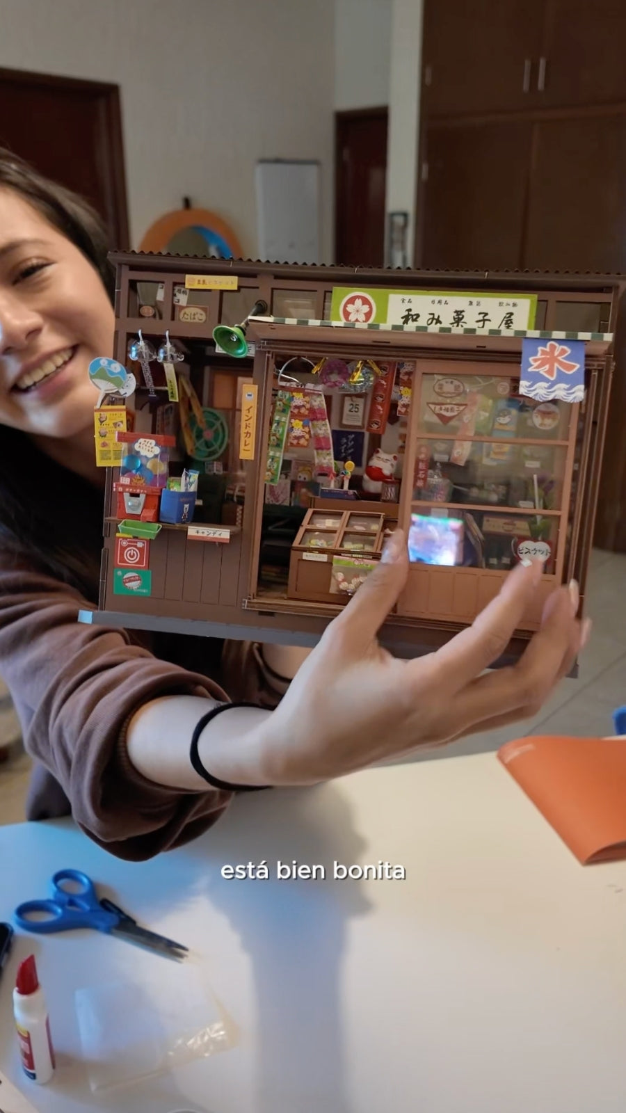Smiling woman holds a finished miniature corner shop during a crafting session