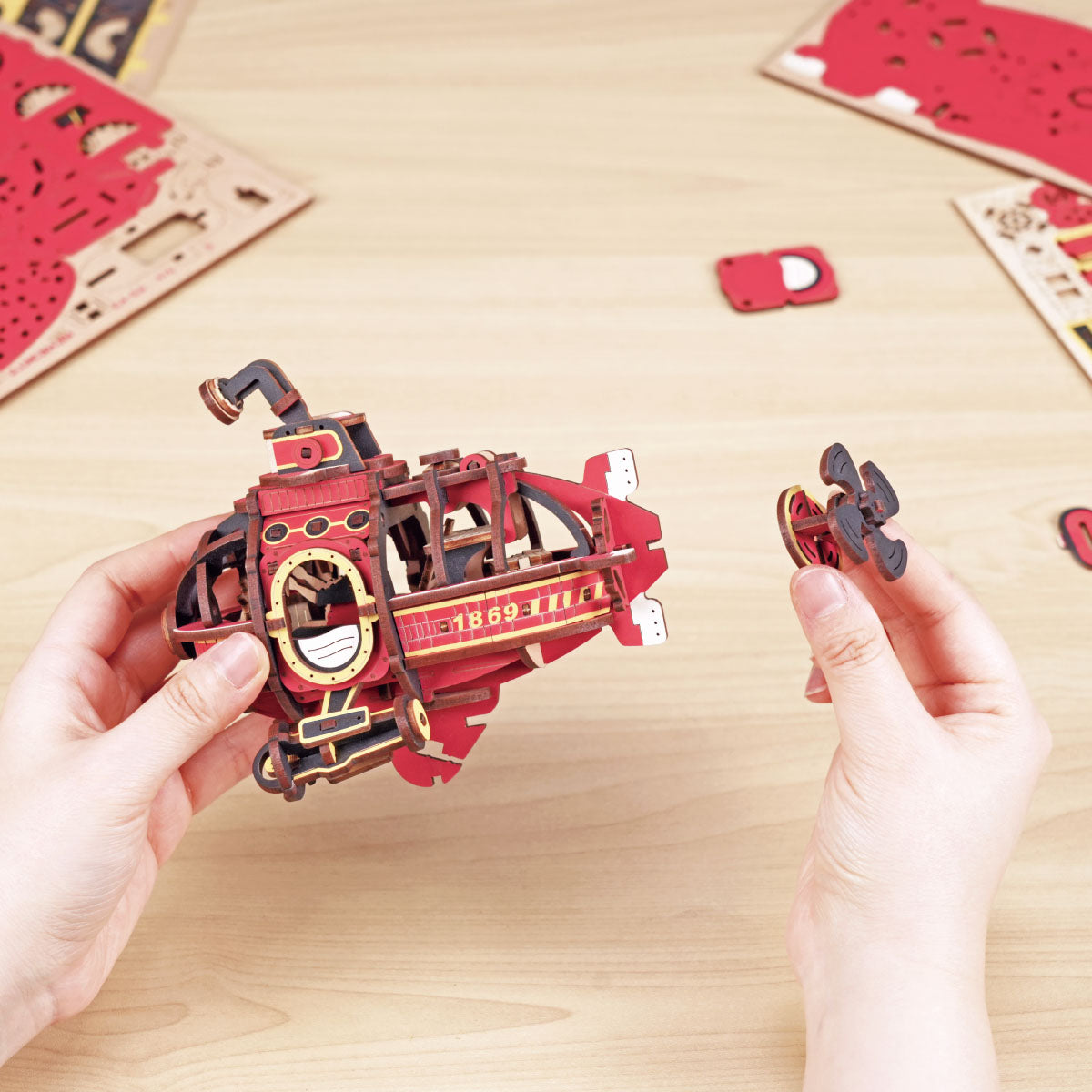 ROKR Deep-Sea Adventurer MCD04 assembly lifestyle showing hands holding the partially built red and gold submarine body beside laser-cut wood sheets
