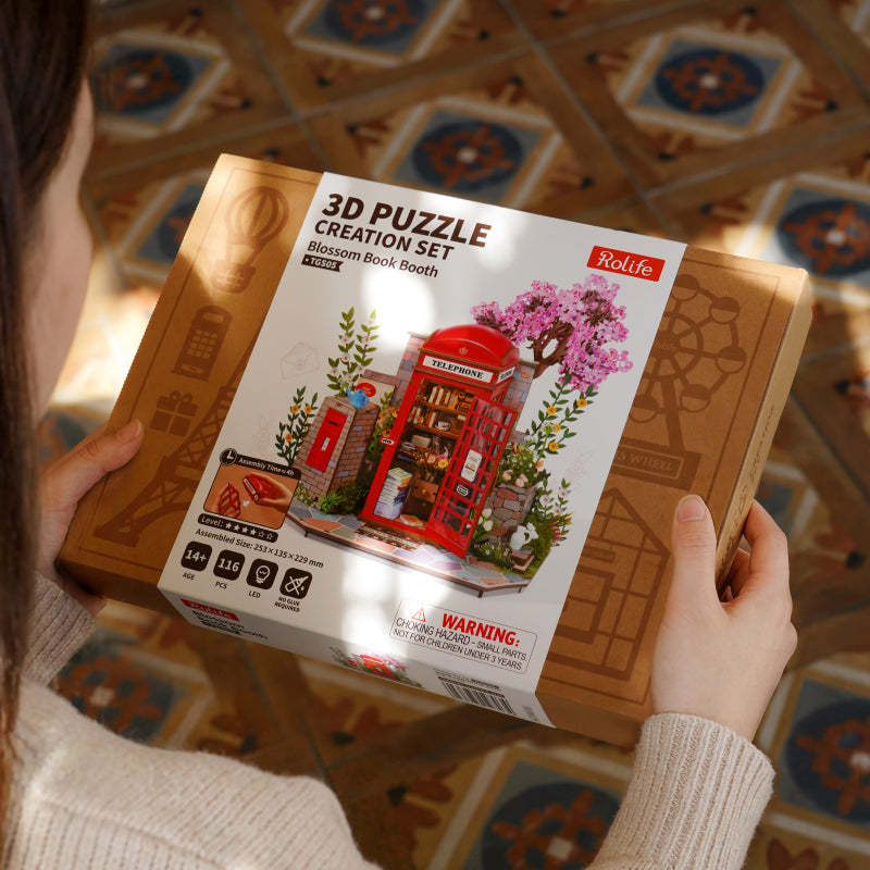 Person holding a boxed Robotime 3D puzzle creation set featuring a red telephone booth design with pink blossom accents