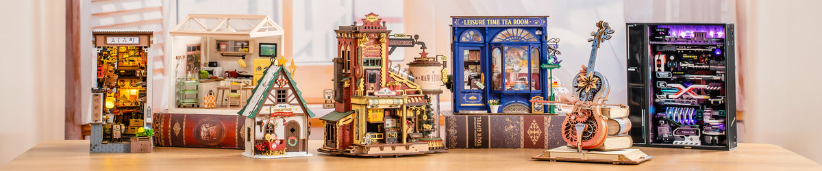 Wide banner showing six Rolife and ROKR book nook and miniature models in a row on a wooden table including a lit Japanese alley, greenhouse, steampunk tower, tea room, violin, and server rack