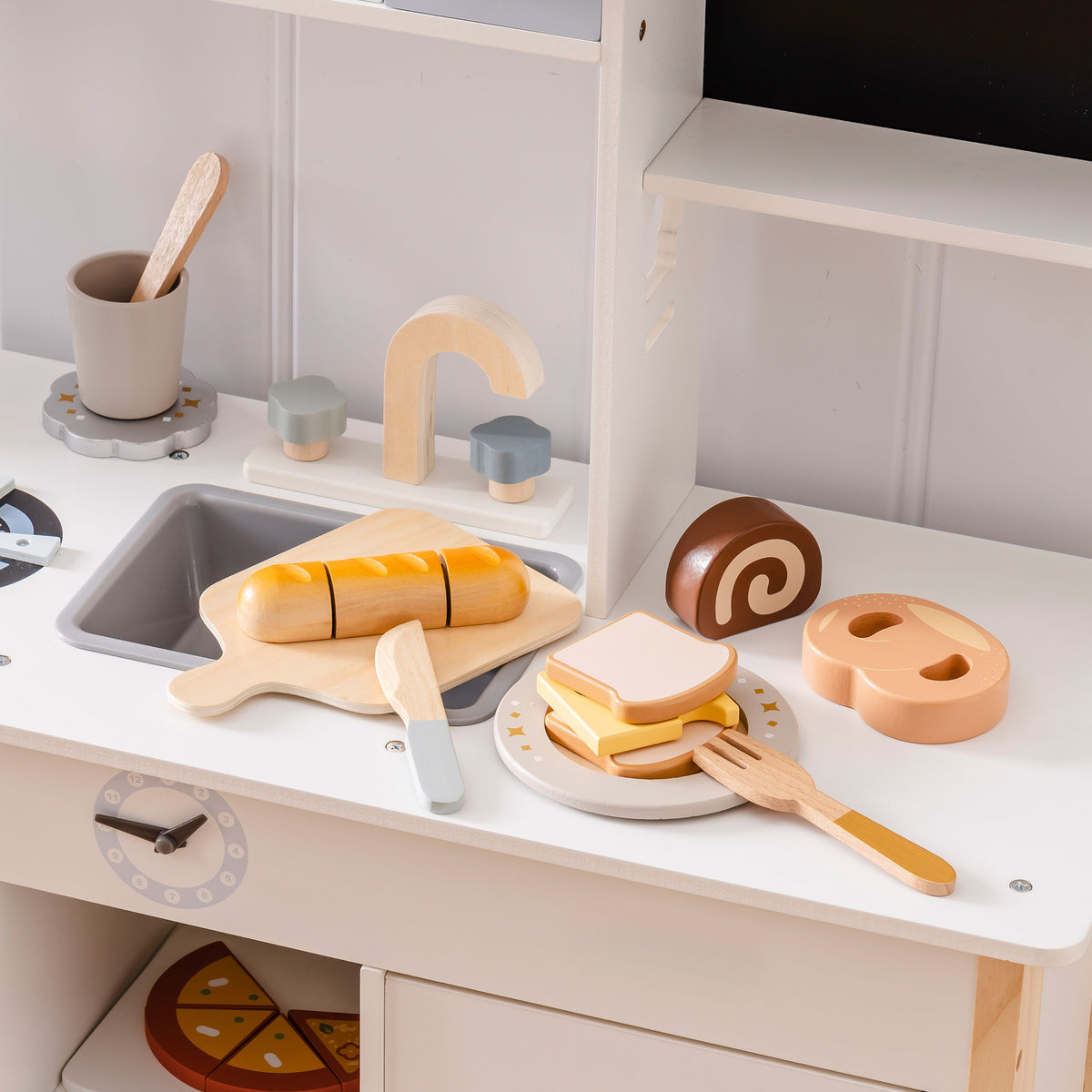 ROBUD wooden bread and dessert cutting set on a white play kitchen counter with baguette on cutting board, toast with cheese on plate, swiss roll and pretzel staged nearby