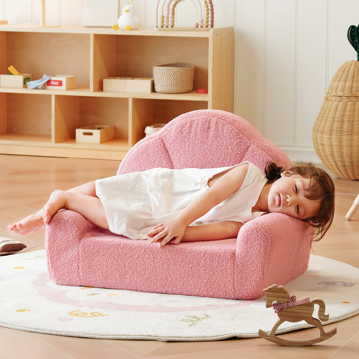 Young girl naps comfortably on the ROBUD pink teddy fleece kids sofa placed on a white rug in a pastel playroom.
