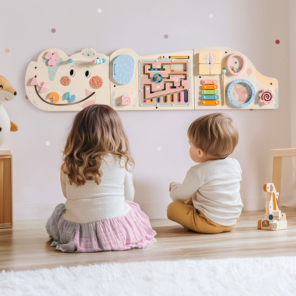 Toddler girl in blue dress reaching up to play with ROBUD Wooden Cream Elephant Busy Board mounted at child height on a light gray wall