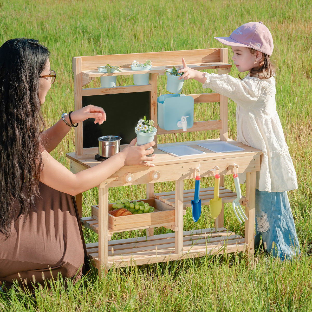 ROBUD Outdoor Garden Mud Kitchen lifestyle scene with mother and girl using the natural wood outdoor mud kitchen together in open grassy field