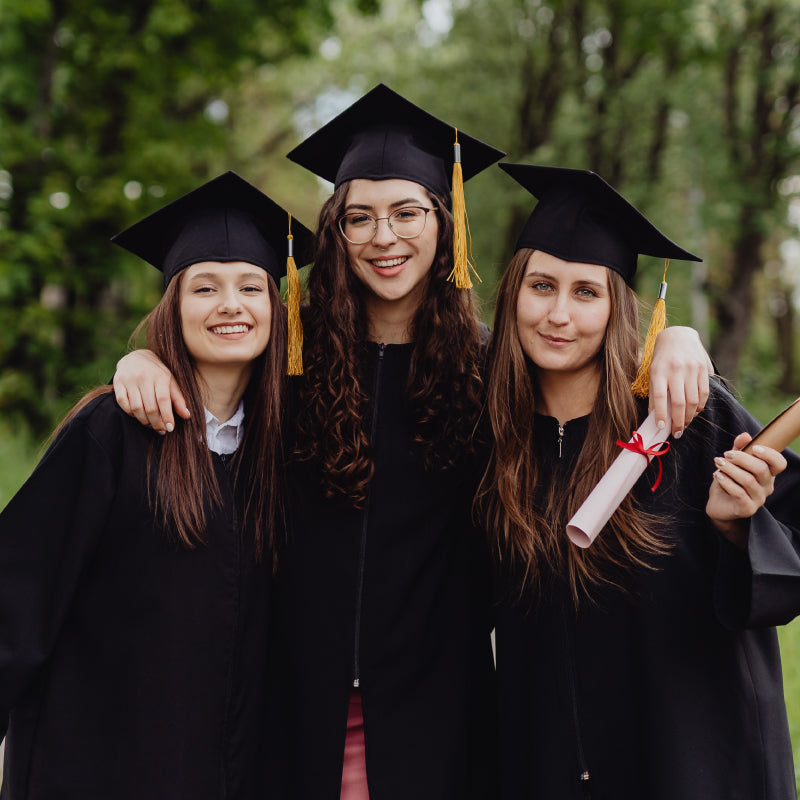 Three young women in black graduation gowns and caps smiling together outdoors among green trees
