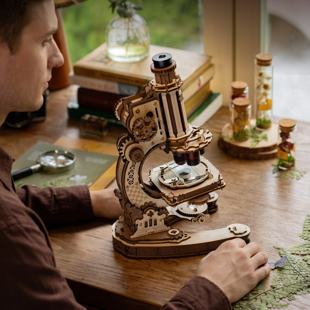 Person studying at a wooden desk with the ROKR wooden microscope in the foreground surrounded by books, plants and glass bottles
