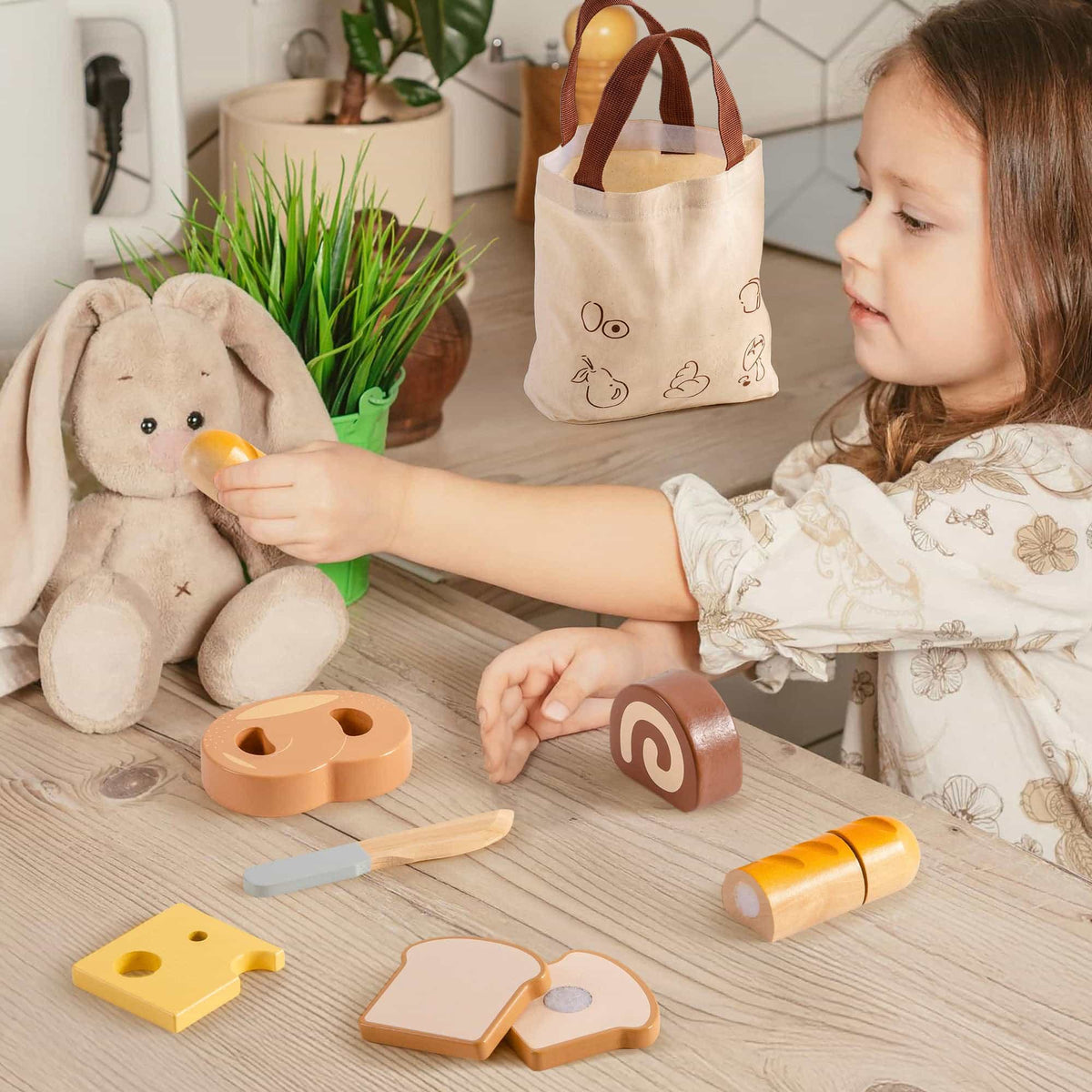 Young girl feeding a toy bunny with a wooden baguette piece from the ROBUD dessert cutting playset while swiss roll, toast, and cheese pieces are on the table
