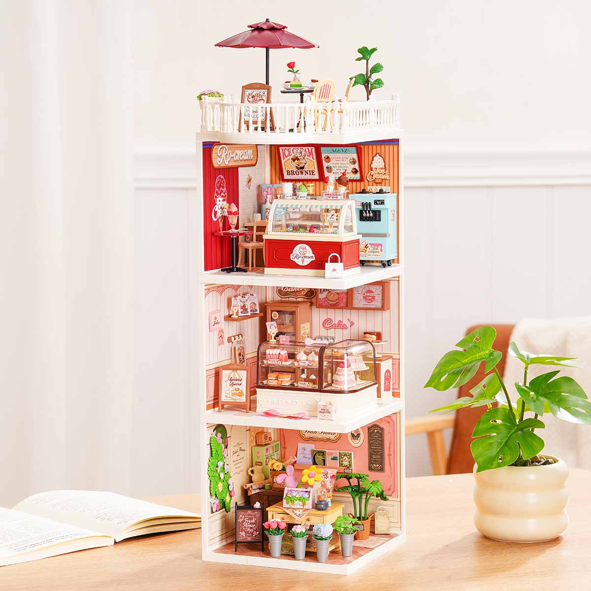 Rolife Super Creator Series 3-in-1 I Shape four-storey miniature tower with rooftop terrace, ice cream shop, cake bakery, and fresh flower shop rooms beside a green plant on a desk.