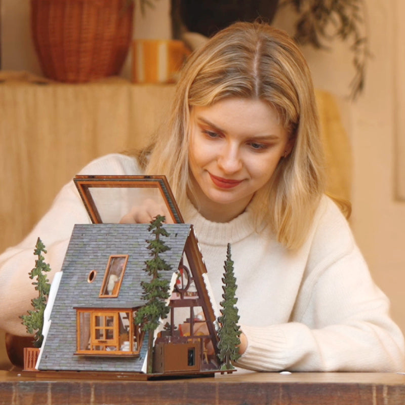Woman assembling a ROKR wooden A frame cabin model kit with miniature pine trees on a wooden tabletop