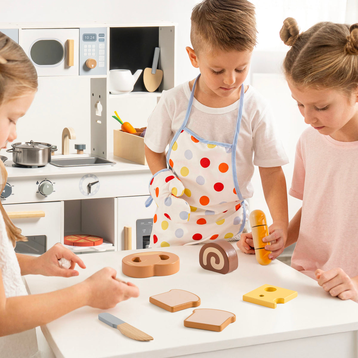 Three children around a white play kitchen pretending to cook with ROBUD wooden bread and dessert cutting pieces including baguette, swiss roll, toast, and cheese