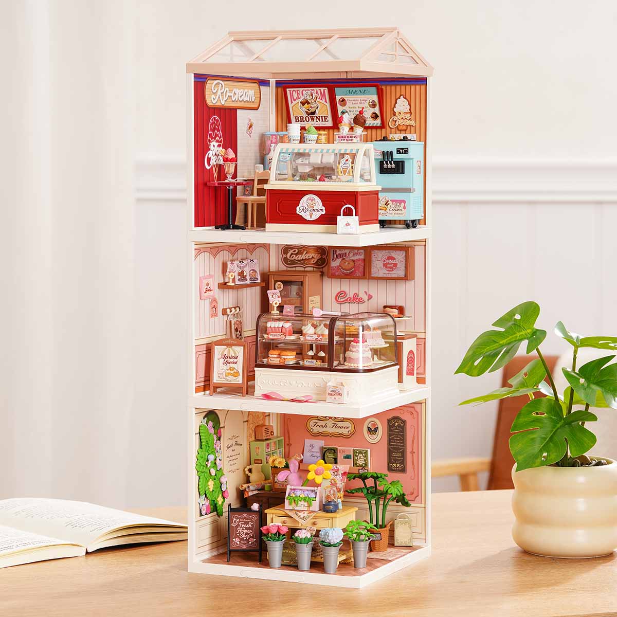 Rolife Super Creator Series 3-in-1 I Shape miniature tower showing an ice cream shop, cake bakery, and flower room with glass roof modules stacked on a wooden surface beside books.