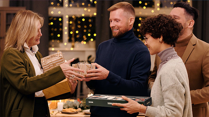 Group of adults exchanging wrapped gifts at a festive holiday gathering under warm string lights, one woman holding a green ROKR branded product box