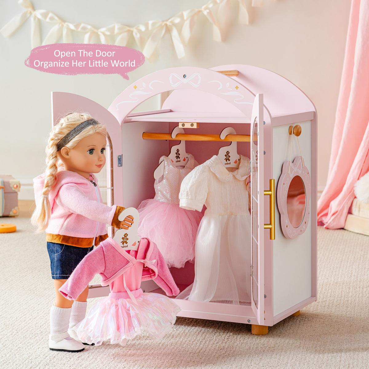 18-inch doll stands beside the open ROBUD pink wooden wardrobe with mini dresses hung inside on white hangers.