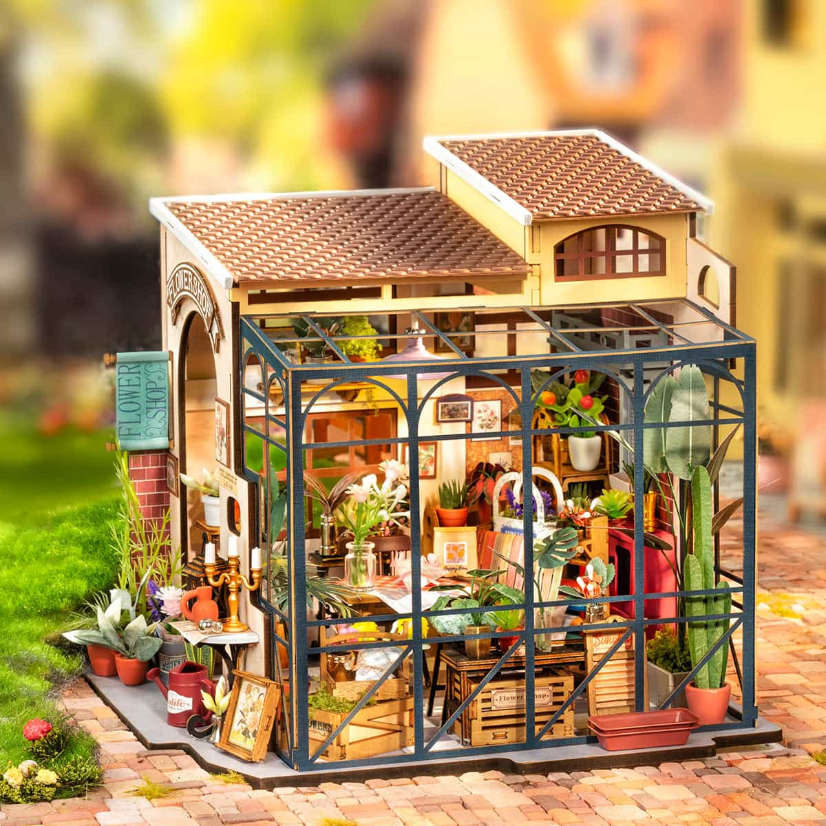 Rolife Emily's Flower Shop DG145 DIY miniature house full exterior view with blue steel greenhouse frame, brick storefront sign, surrounding potted plants and red watering can on table