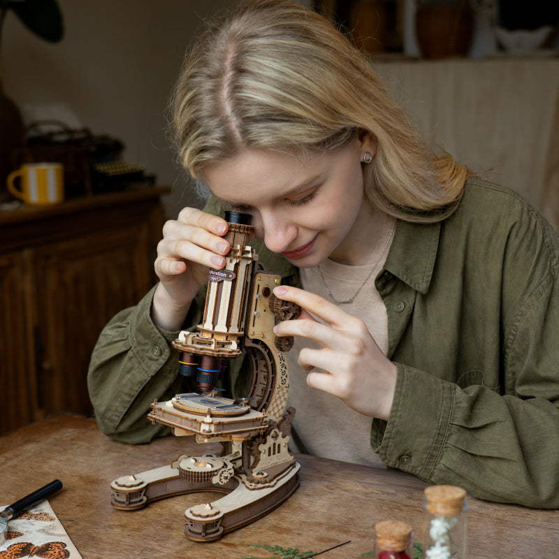 Woman looking through the eyepiece of a ROKR wooden microscope model kit while seated at a wooden table