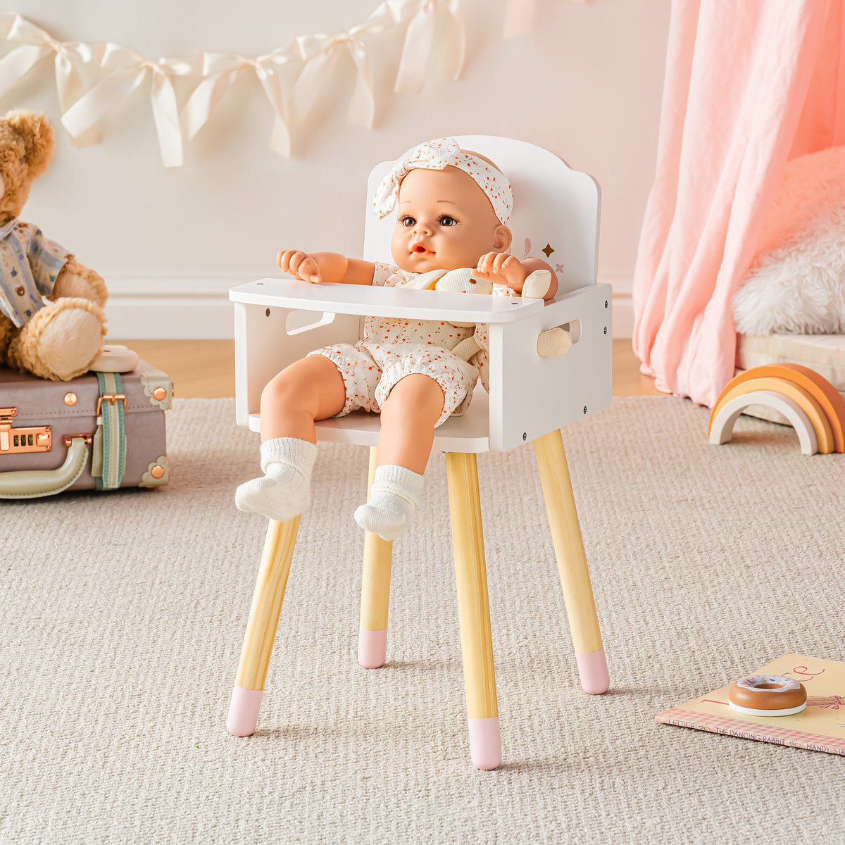 Baby doll in bonnet seated alone in the ROBUD white doll high chair with pink accents on a neutral beige rug.