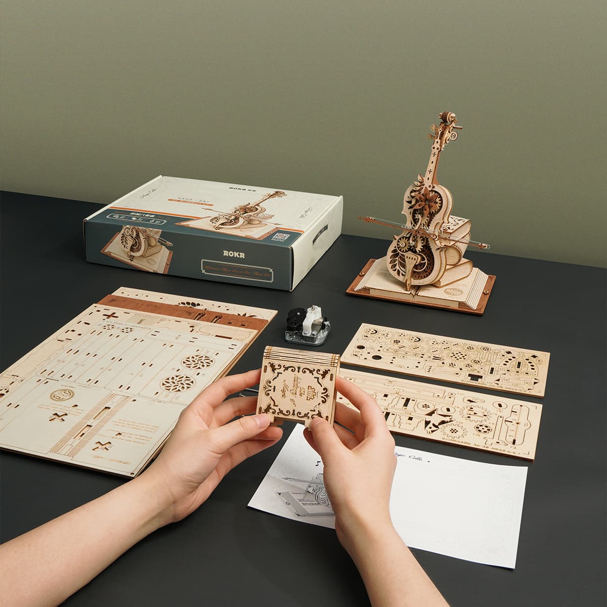 Hands sorting laser-cut sheets beside assembled ROKR Magic Cello music box model and its retail packaging on dark table