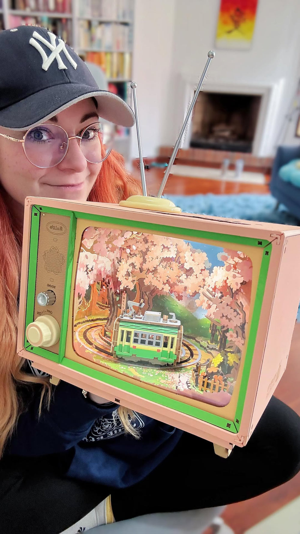 Fan holding a completed Robotime ROKR wooden mechanical TV puzzle with a cherry blossom tram diorama inside, in a cozy home setting