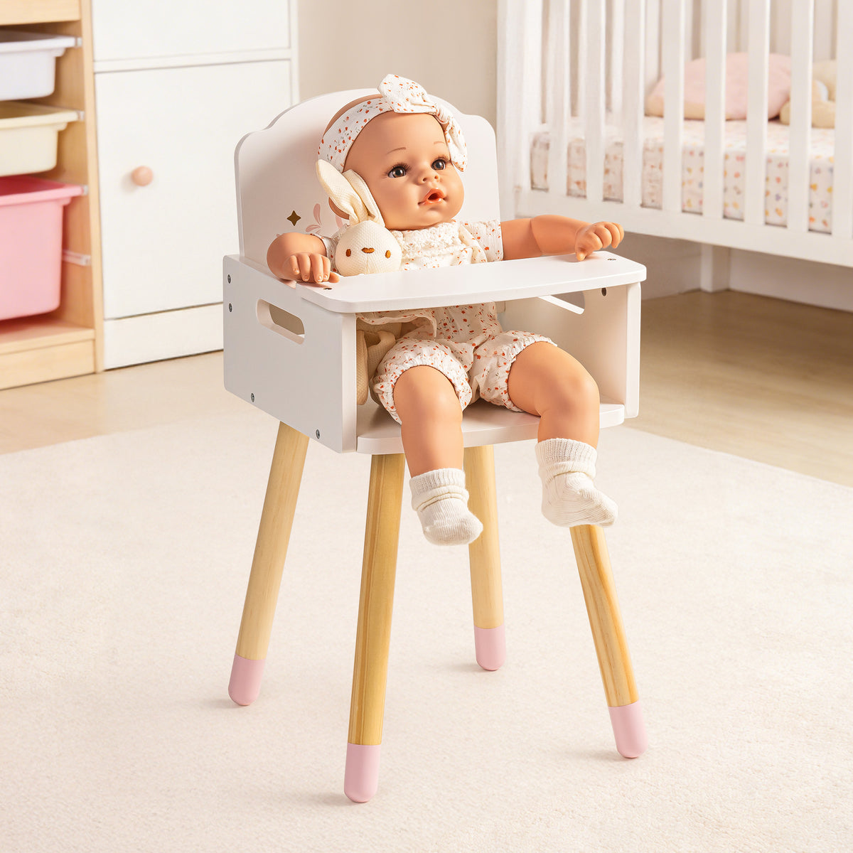 Baby doll seated in ROBUD white wooden high chair with pink-tipped legs shown in a nursery with crib in background.