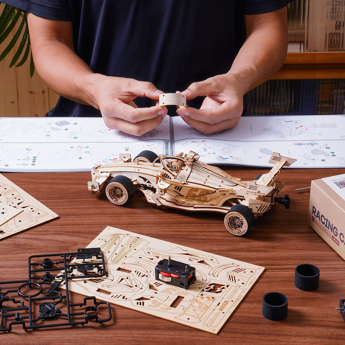 Hands assemble the ROKR racing car kit on instructions with wooden parts spread across a table.