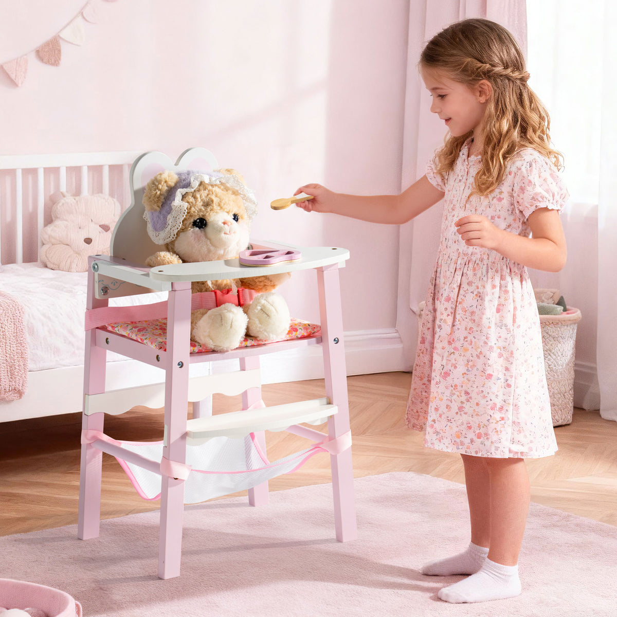 Girl in a floral dress feeds a stuffed bear seated in the ROBUD pink wooden doll high chair in a styled pastel nursery room.