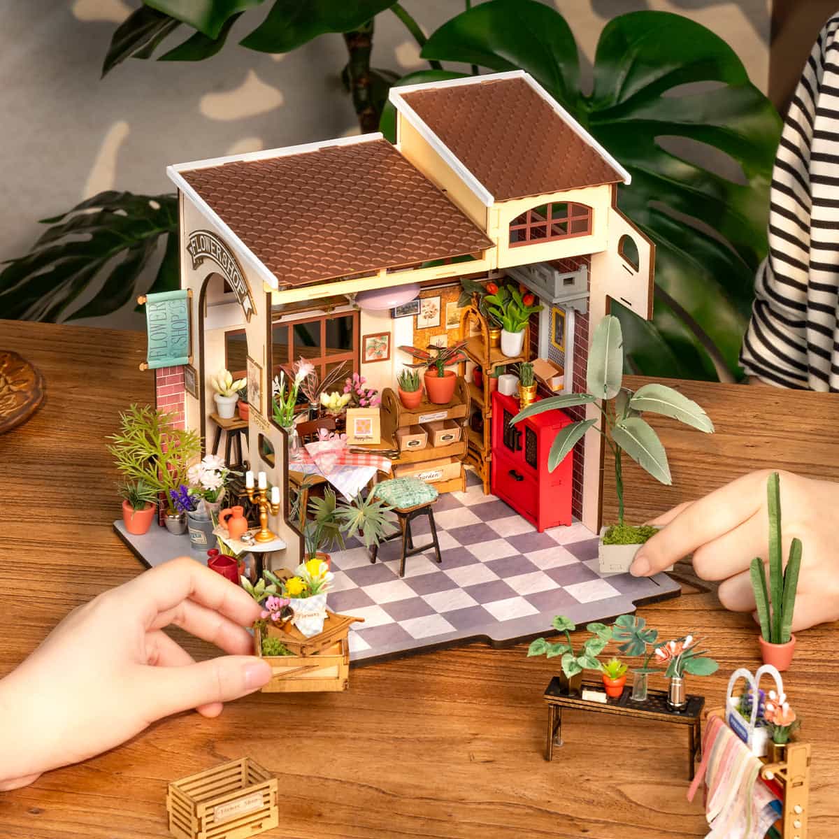 Rolife Emily's Flower Shop DG145 miniature house seen from the front on a wooden table as hands place small furniture pieces, with tropical plants beside it