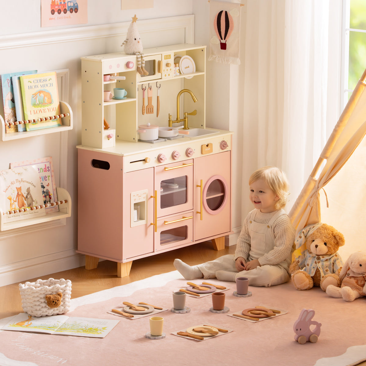 Toddler girl sitting beside the ROBUD Glowwood pink play kitchen in a warm nursery with a teepee in the background.