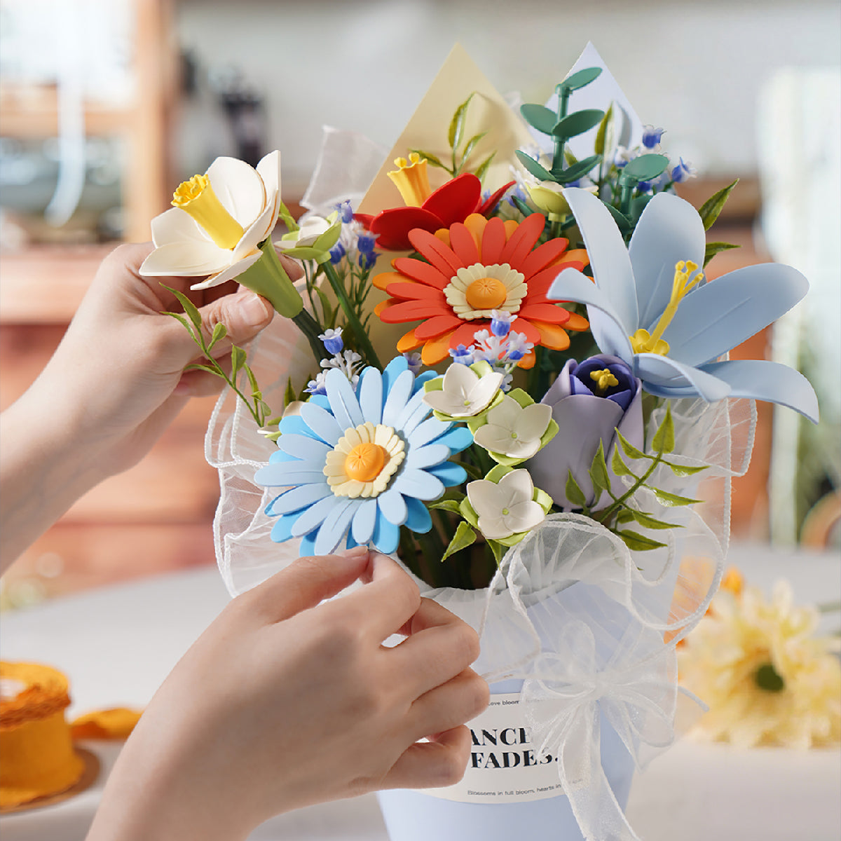 Hands arranging a Rowood foam bouquet of orange gerberas, blue daisies and white lilies in organza ribbon