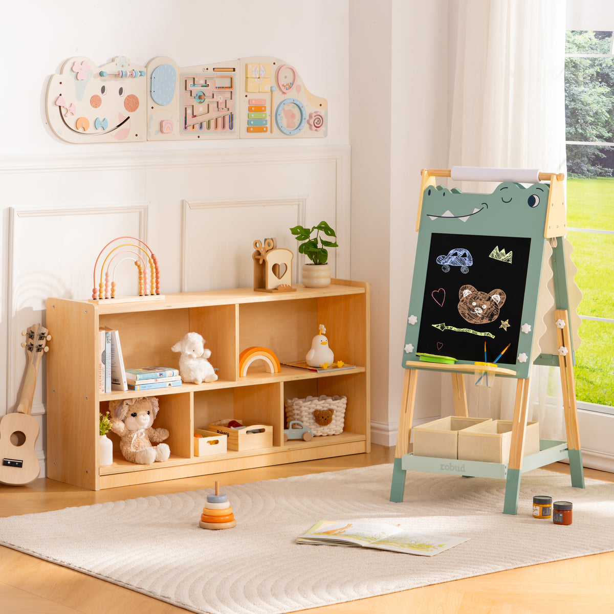 ROBUD dino art easel with chalk illustrations displayed beside wooden bookcase in an airy pastel playroom corner.