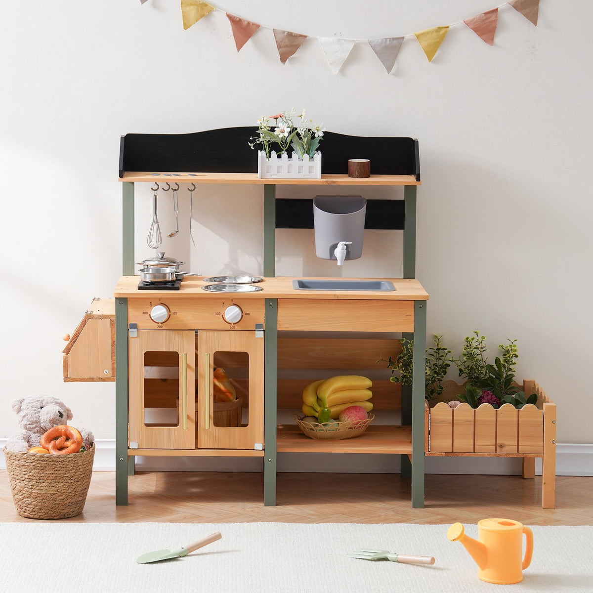 ROBUD outdoor wooden mud kitchen styled indoors against a white wall with stainless steel pots, hanging utensils, chalkboard panel, planter box with greenery, and fruit basket below