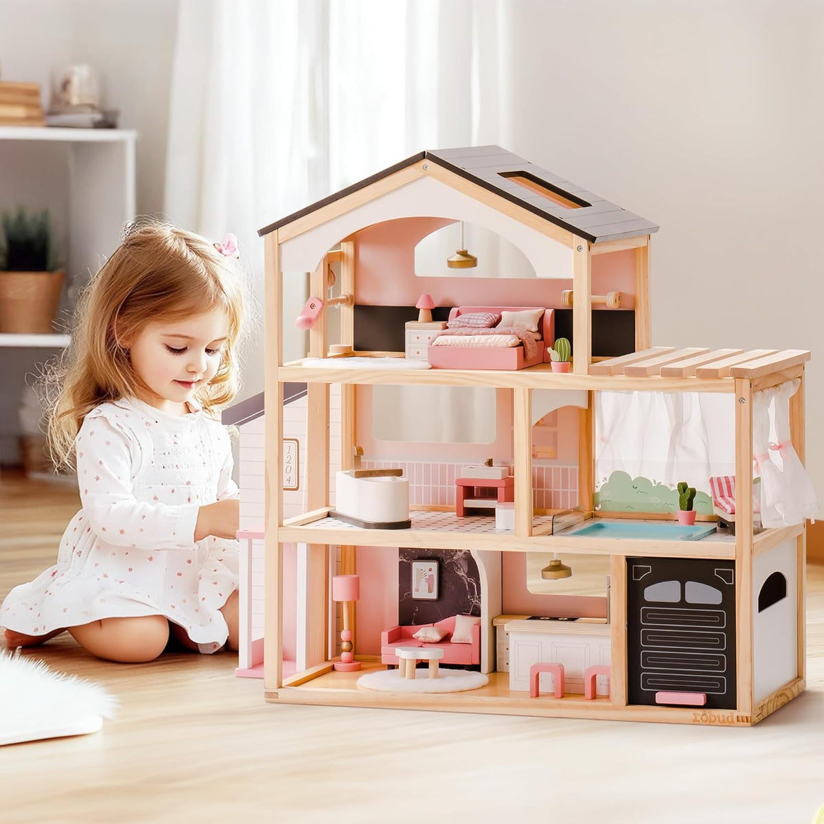 Robud three-story wooden dollhouse with a girl kneeling beside it interacting with the open-plan pink-furnished floors including bedroom, bathroom and kitchen