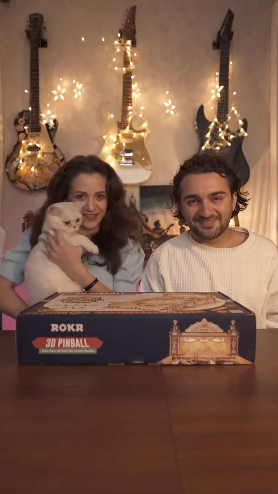 Smiling couple seated behind a ROKR model kit box beneath guitars and string lights