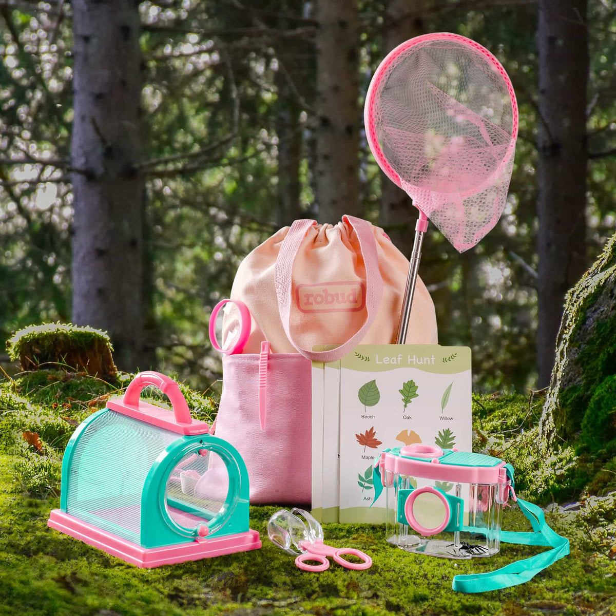 ROBUD outdoor kids explorer kit in pink laid on mossy forest ground showing bug catcher cage, magnifying jar, pink butterfly net, leaf hunt guide card, and pink drawstring bag