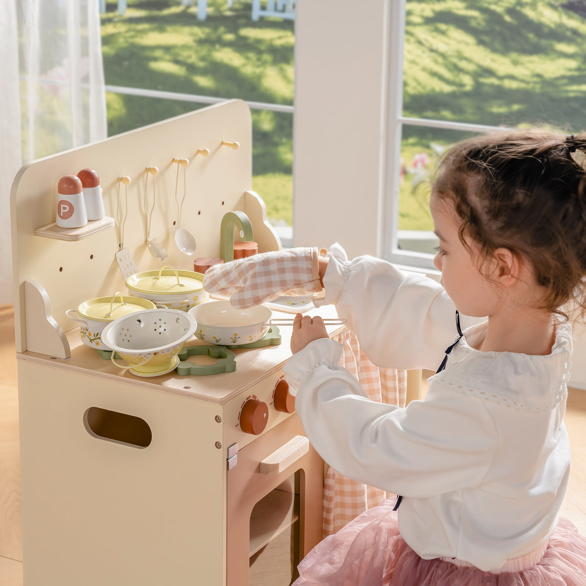 Smiling girl standing at wooden play kitchen using ROBUD Tinplate Pretend Cooking Utensils pink series with microwave and sink in view
