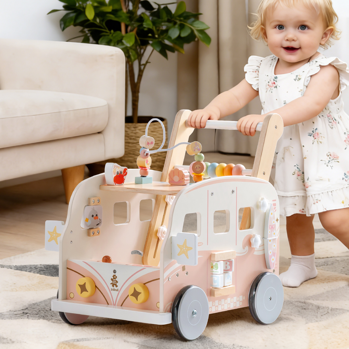 Toddler girl in a white dress pushes the ROBUD pink vacation bus walker across a bright living room with plants.