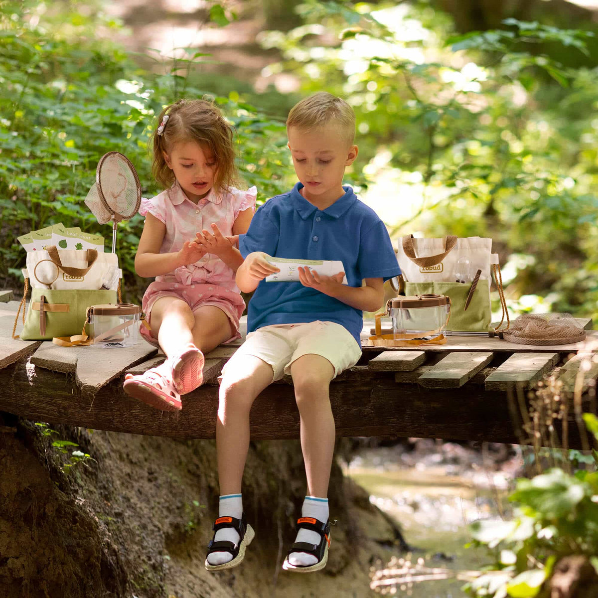 Two children sitting on a wooden bridge in the forest reading a guide card with ROBUD explorer kit tools spread around them
