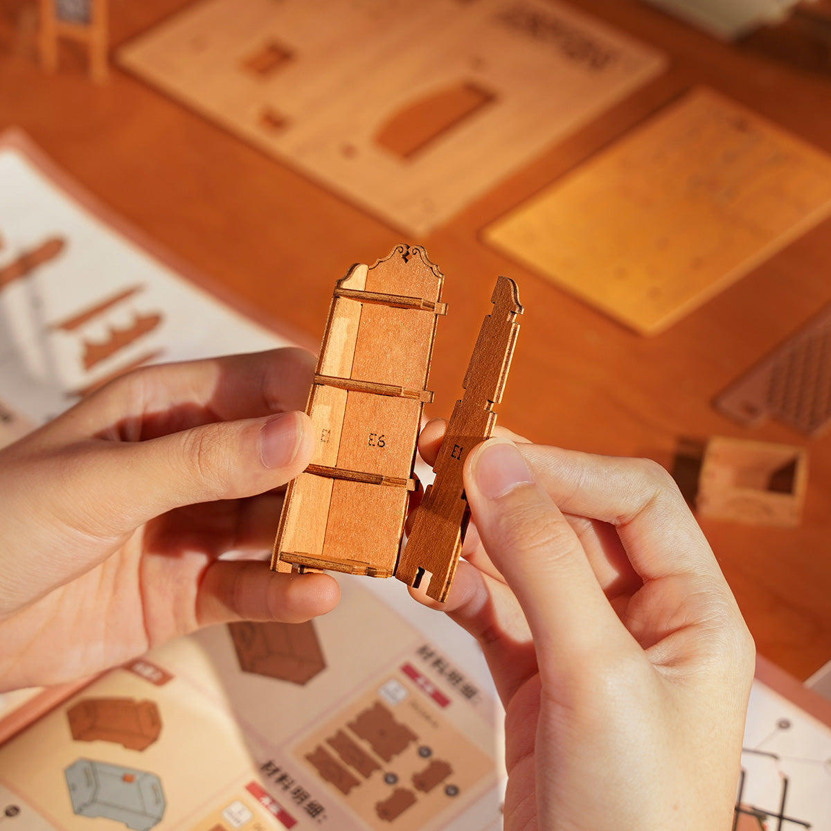 Hands holding a small wooden furniture piece during Rolife Corner Bookstore assembly with laser-cut parts and instruction sheet spread on a desk