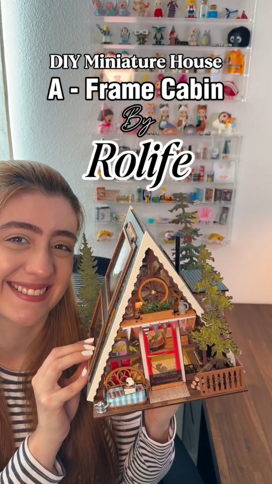 Woman holding an A frame cabin miniature with flowers, furniture and a steep wooden roof