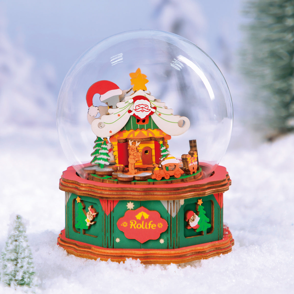 Rolife Christmas Snow Globe AM46 displayed on snow in outdoor winter setting with pine trees showing full illuminated festive globe scene