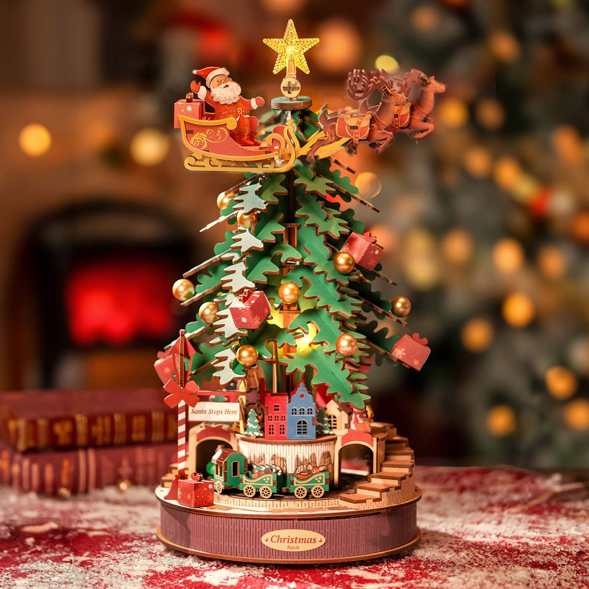 Rolife Christmas Melody Tree AMS01 holiday table setting with the lit rotating tree displayed beside an open book and festive red backdrop