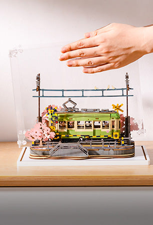 Hand lowering an acrylic display cover over a finished wooden cherry blossom tram miniature placed on a light wooden surface